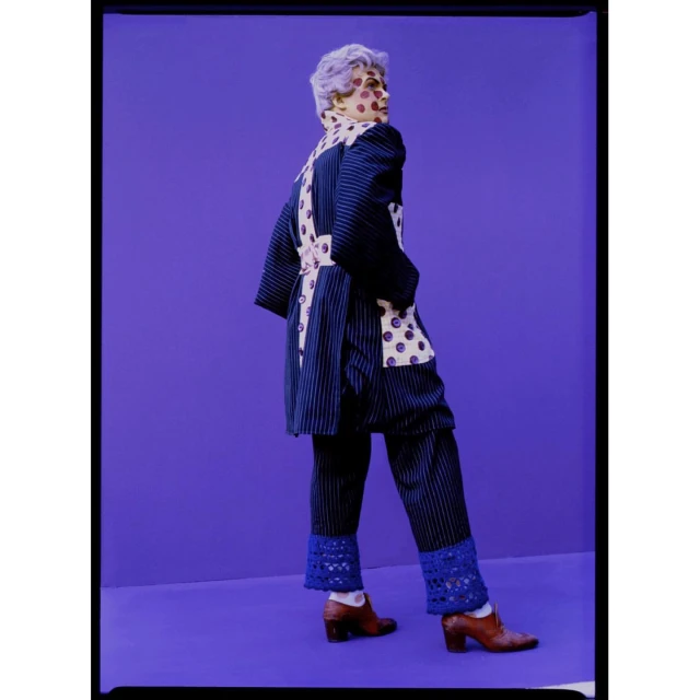 Adult female in a blue pinstripe coat and pants ensemble poses against a purple background, complemented by brown heeled shoes and short hair.