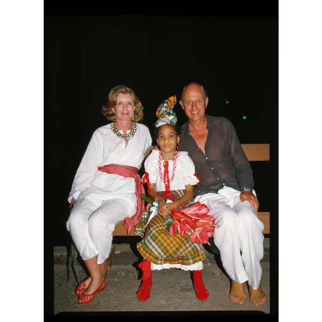 Woman, girl and man pose on bench in formal wear. Adult female in white dress with red sash, child in dress with red boots.