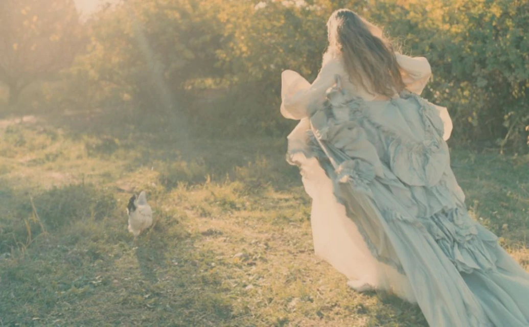 A woman in a ruffled dress runs through a grassy field toward trees in the background, with a chicken in the foreground.