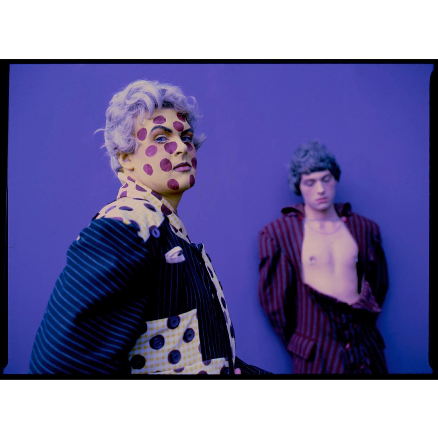 Two adult males with painted faces and striped jackets, posing in front of a flat purple background, in a vibrant, eye-level fashion shot.