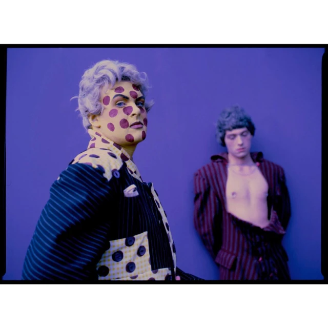 Two adult males with painted faces and striped jackets, posing in front of a flat purple background, in a vibrant, eye-level fashion shot.