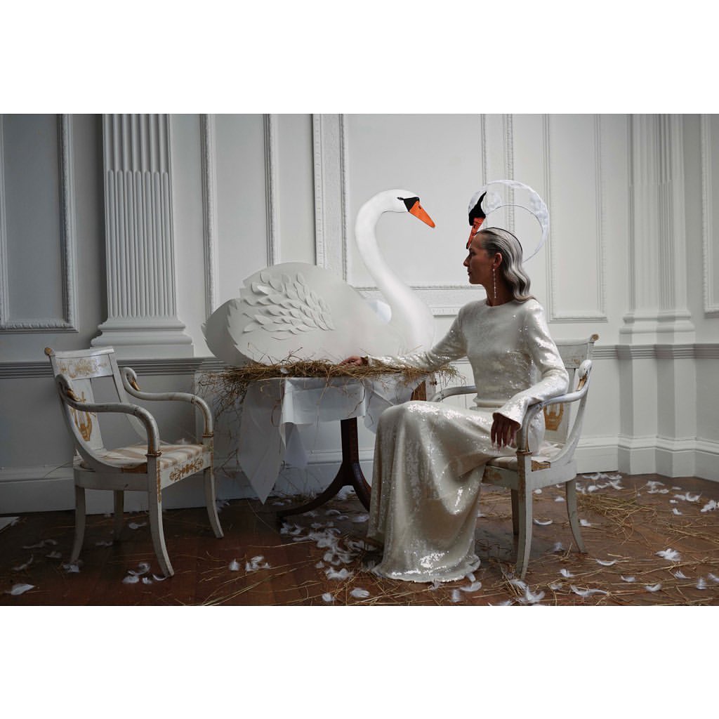 Elegant woman in a gown seated next to a large paper swan, bathed in soft light within an ornate interior, evoking a vintage fashion aesthetic.