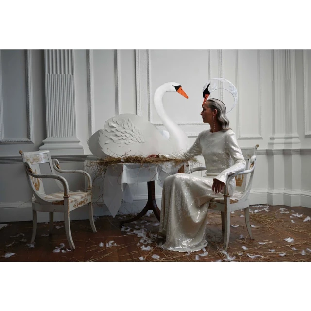 Elegant woman in a gown seated next to a large paper swan, bathed in soft light within an ornate interior, evoking a vintage fashion aesthetic.