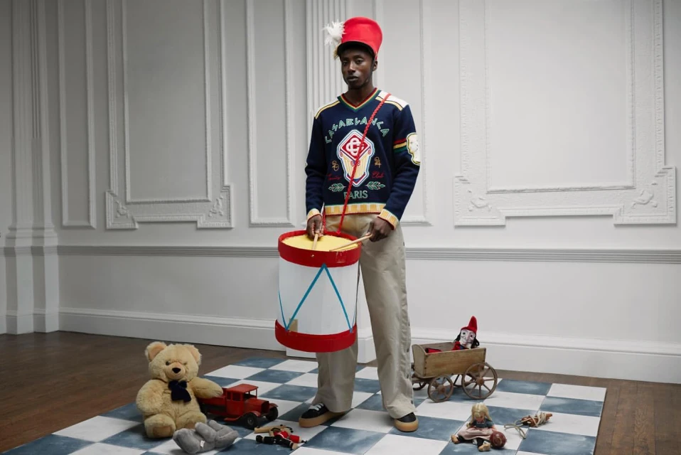 An adult male model poses indoors wearing a blue sweater, red hat, and holding a white drum, surrounded by various toys on a checkered mat.