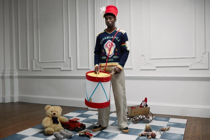 An adult male model poses indoors wearing a blue sweater, red hat, and holding a white drum, surrounded by various toys on a checkered mat.