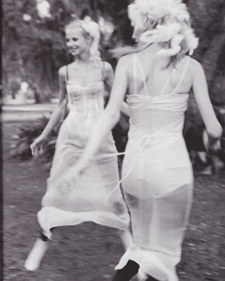 Two adult women in white dresses pose outdoors with blurred motion, soft lighting, and a vintage, black-and-white aesthetic.