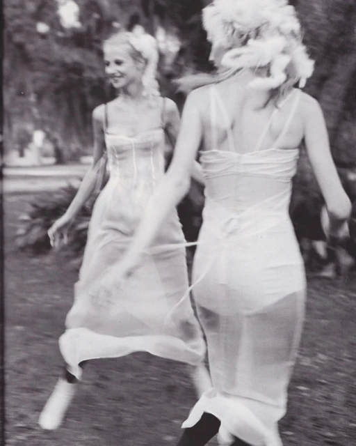 Two adult women in white dresses pose outdoors with blurred motion, soft lighting, and a vintage, black-and-white aesthetic.