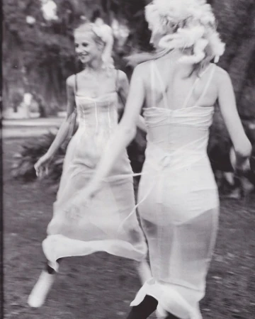 Two adult women in white dresses pose outdoors with blurred motion, soft lighting, and a vintage, black-and-white aesthetic.