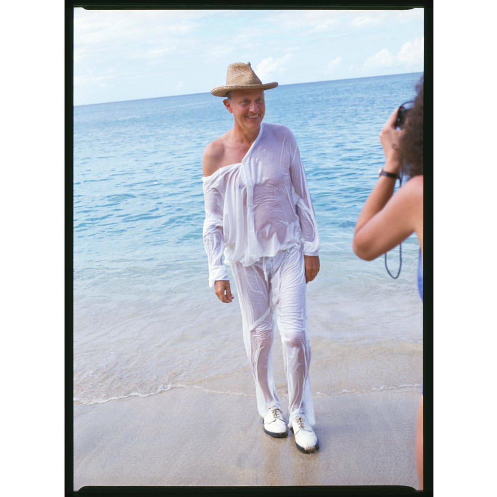 A man in wet white clothing and a straw hat standing on a beach with waves, being photographed by a person holding a camera.