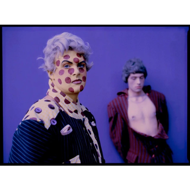 Two people in surreal fashion. One with polka dot makeup and a striped jacket stands in front of another in an open jacket.
