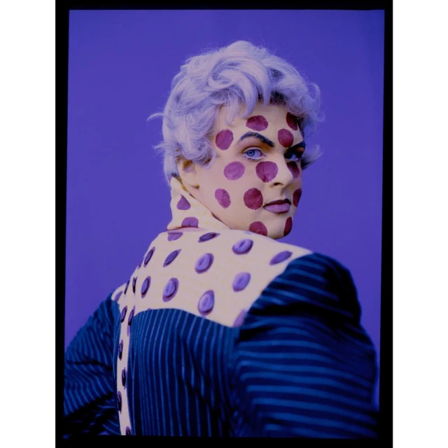 Unique studio portrait of a person with white hair, bold makeup, and a blue-striped jacket with a polka-dot collar, set against a purple backdrop.