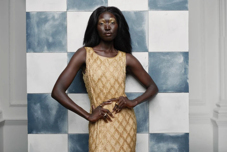 Adult woman poses in a gold dress in a studio. She stands in front of blue and white tiles, creating a stylish and elegant portrait.