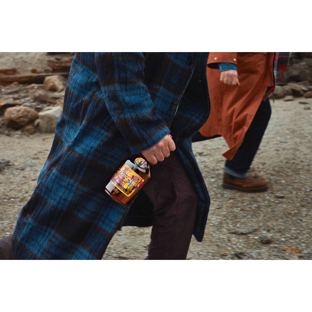 Two people walking on a rocky beach, one carrying a bottle of hard seltzer in a blue plaid coat, showcasing a lifestyle and beverage product.