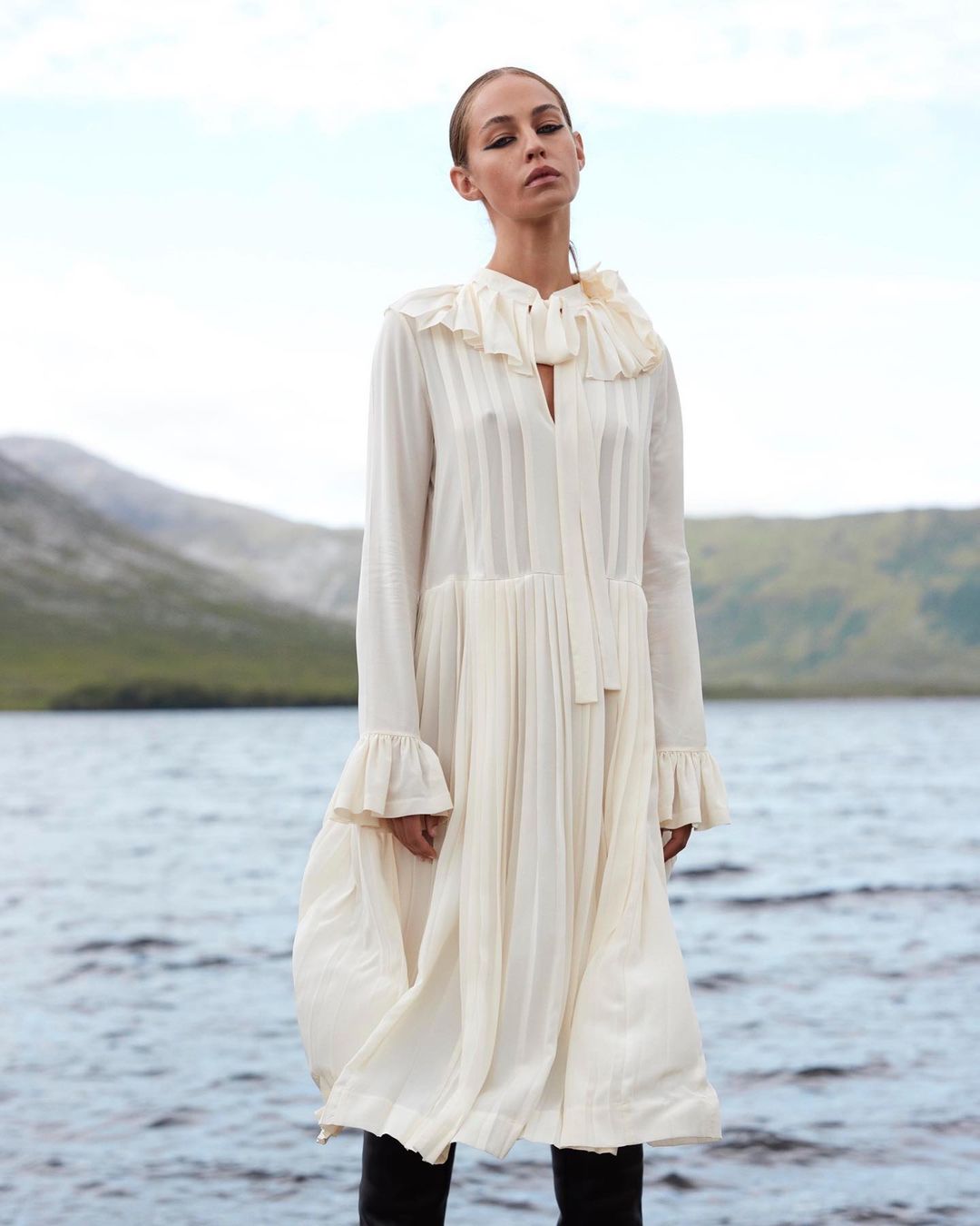 A woman in a white dress stands near a body of water, with mountains visible in the background, in an outdoor setting.