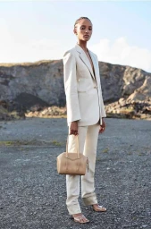 Model in a cream suit holding a handbag standing on a rocky terrain under a clear sky