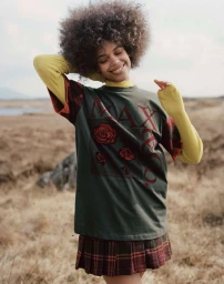 Smiling woman with voluminous afro hair wearing a green graphic t-shirt over yellow sleeves and a plaid skirt outdoors.
