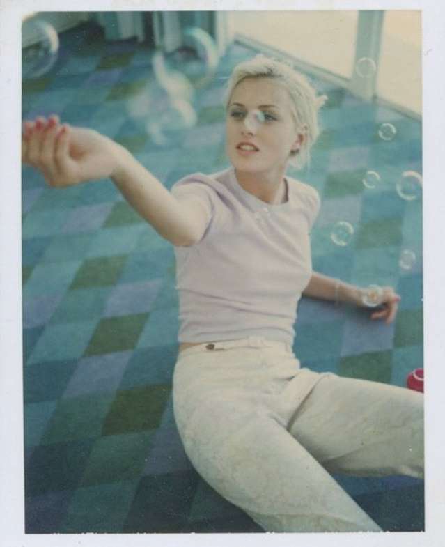Blonde woman wearing lavender t-shirt and light trousers blowing bubbles on a geometric patterned carpet by Perry Ogden