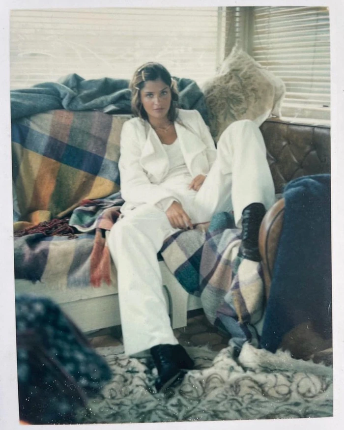 A woman in a white suit and dark boots seated on a plaid-covered sofa by a window, captured by Perry Ogden.