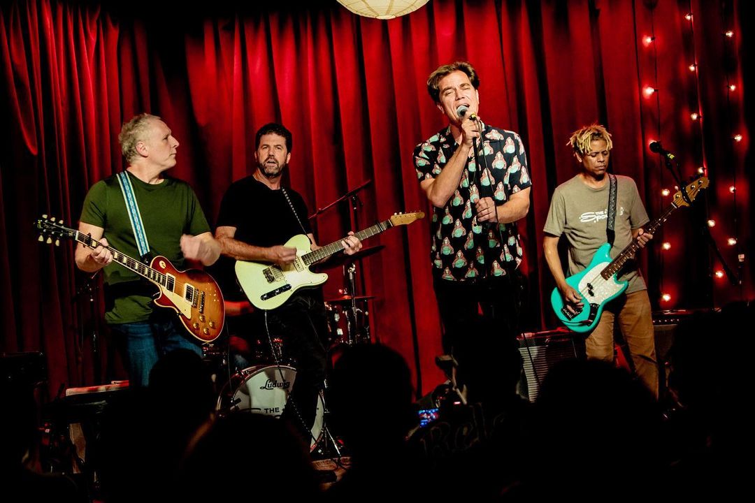 Live music performance features four musicians playing guitars and singing on stage with red curtains in the background, viewed by a crowd.