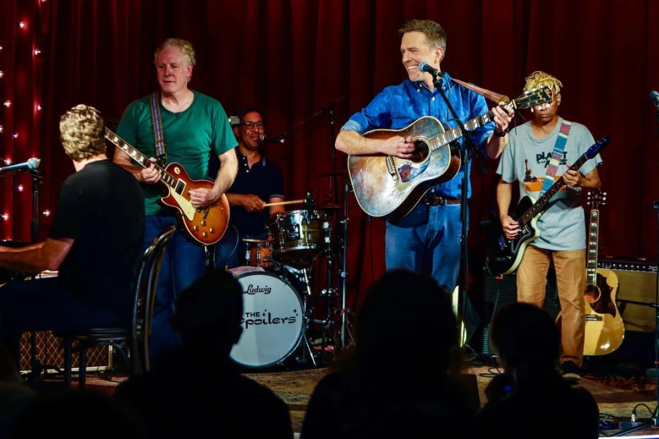 Live music performance featuring four musicians playing guitars and drums on a stage with a red curtain backdrop and silhouetted audience.