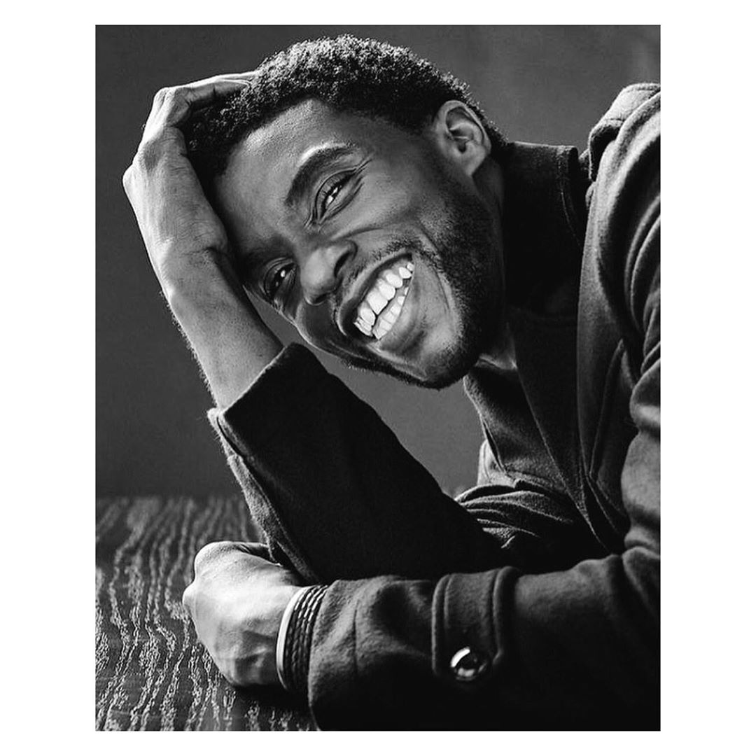 Black and white portrait of a smiling adult male with dark hair resting his head on his hand while posing indoors.