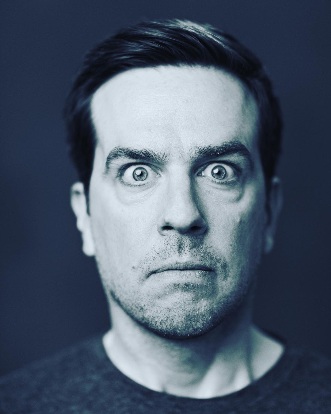 Close-up portrait of a man with dark hair and eyes wide open against a dark blue background, creating a striking and expressive image.