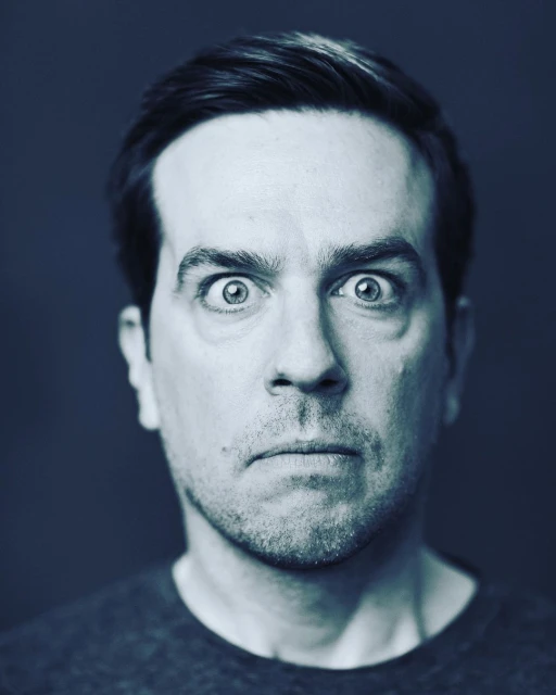 Close-up portrait of a man with dark hair and eyes wide open against a dark blue background, creating a striking and expressive image.