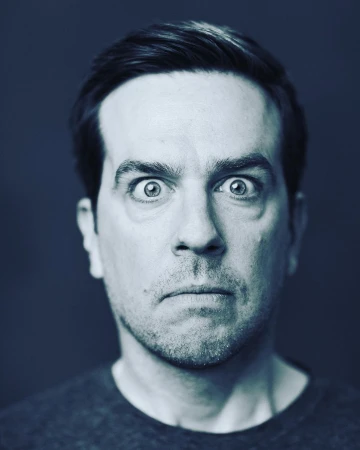 Close-up portrait of a man with dark hair and eyes wide open against a dark blue background, creating a striking and expressive image.
