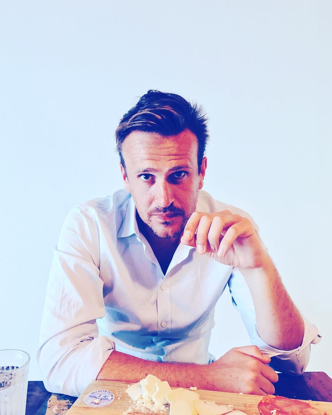 Adult male in white shirt sits indoors at a wooden table with a cheese board and glass, gazing at the camera in a medium shot.
