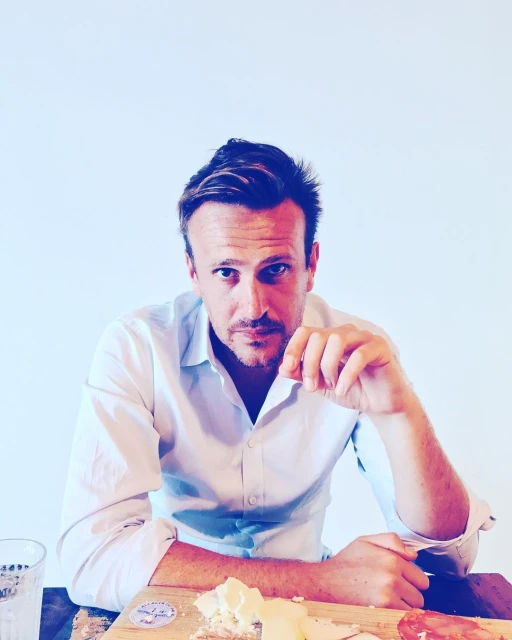 Adult male in white shirt sits indoors at a wooden table with a cheese board and glass, gazing at the camera in a medium shot.