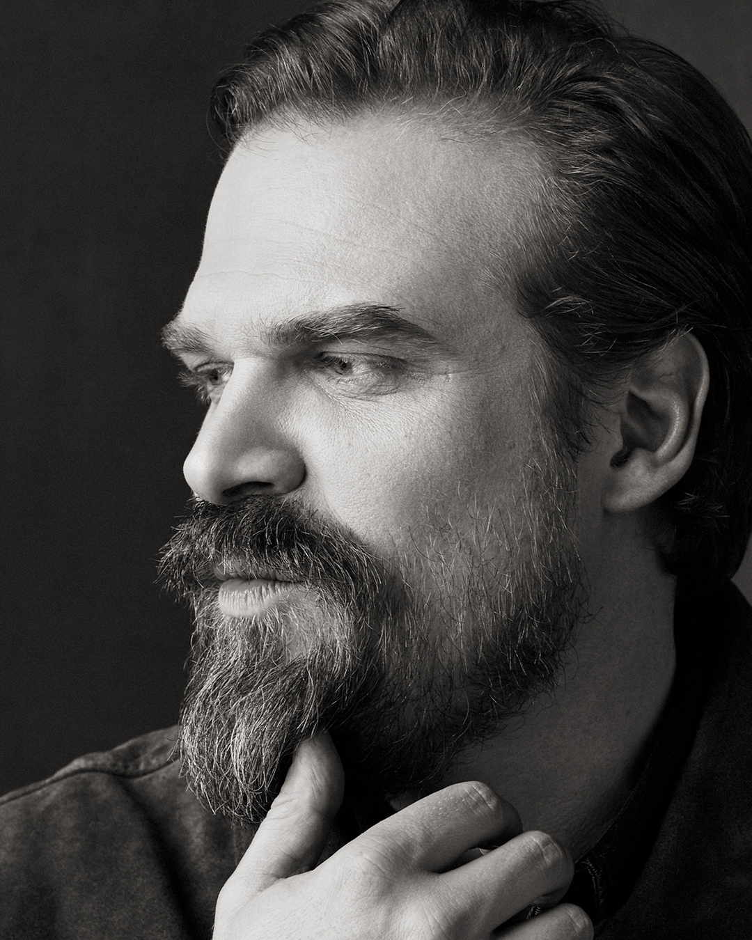 Monochrome portrait of an adult male with a beard and a dark shirt. The side-lit studio shot emphasizes textures and shadows on his face.