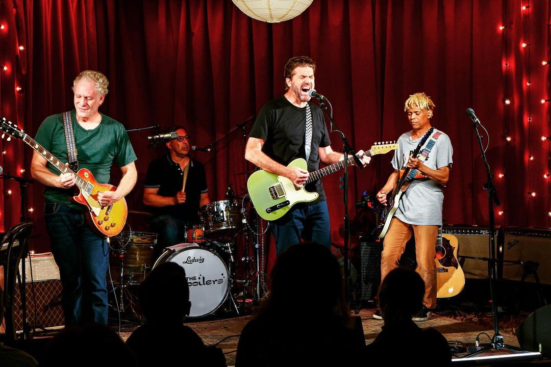 Four musicians perform live on stage with guitars and drums in a concert setting with red curtains and audience silhouettes.