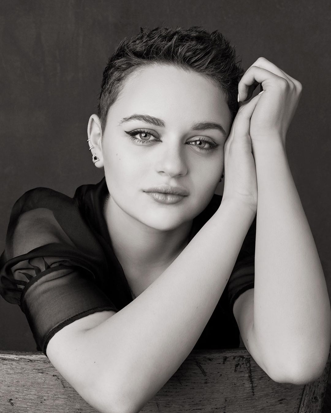 Black and white portrait of a young woman with a short haircut, wearing a sheer top, posing indoors with hands near her face.