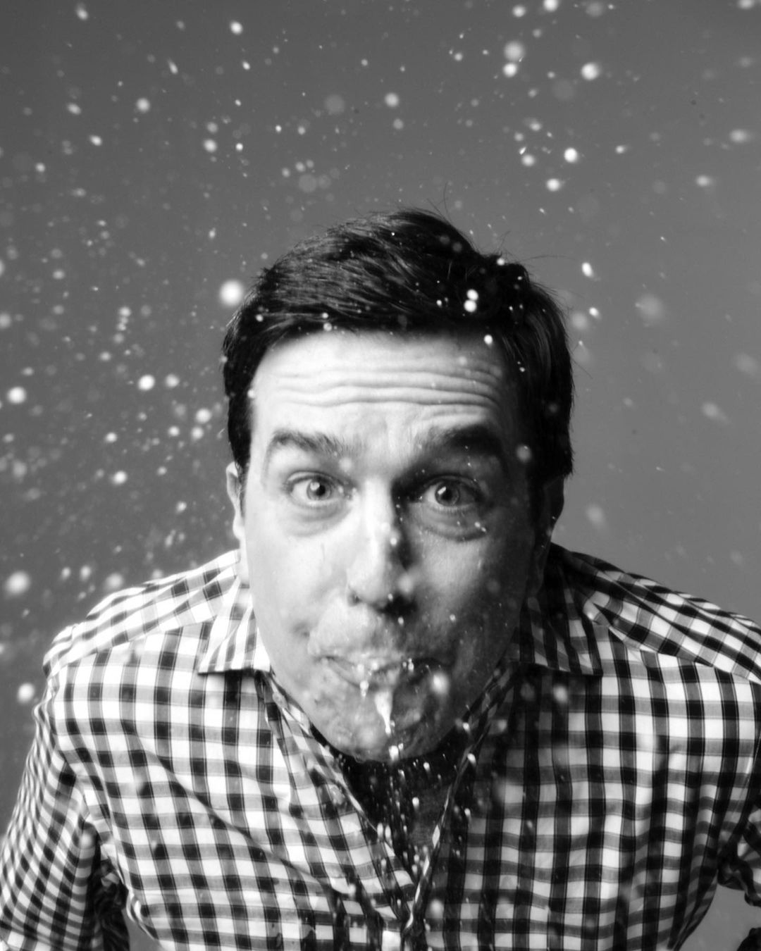 Black and white portrait of a man with a surprised expression, spitting a stream of liquid into the air, wearing a gingham shirt.