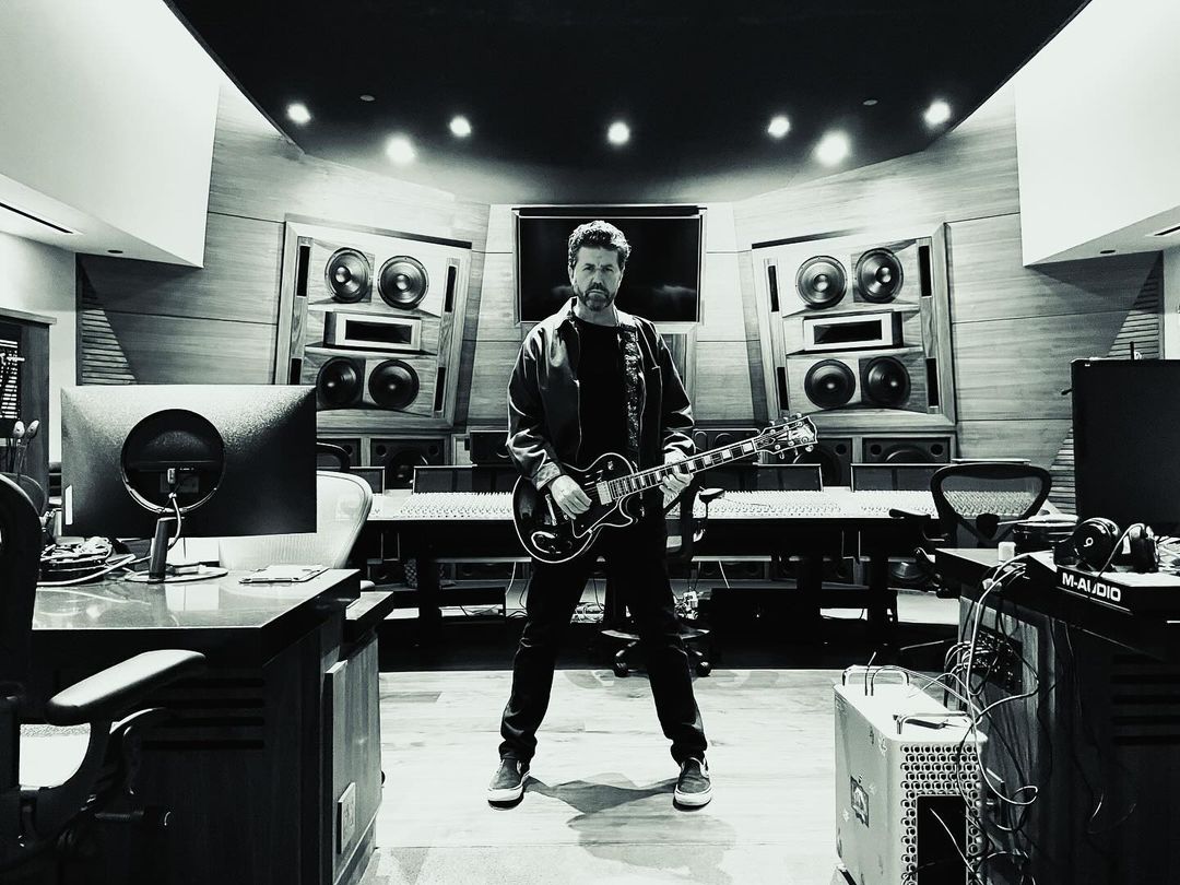 Monochrome shot in a music studio with a man standing, holding an electric guitar, surrounded by audio equipment and soundproofing.