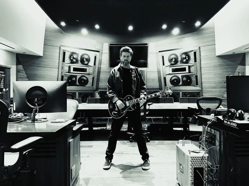 Monochrome shot in a music studio with a man standing, holding an electric guitar, surrounded by audio equipment and soundproofing.