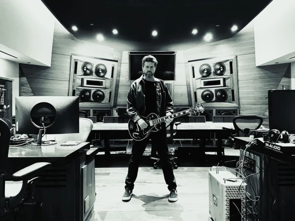 Monochrome shot in a music studio with a man standing, holding an electric guitar, surrounded by audio equipment and soundproofing.