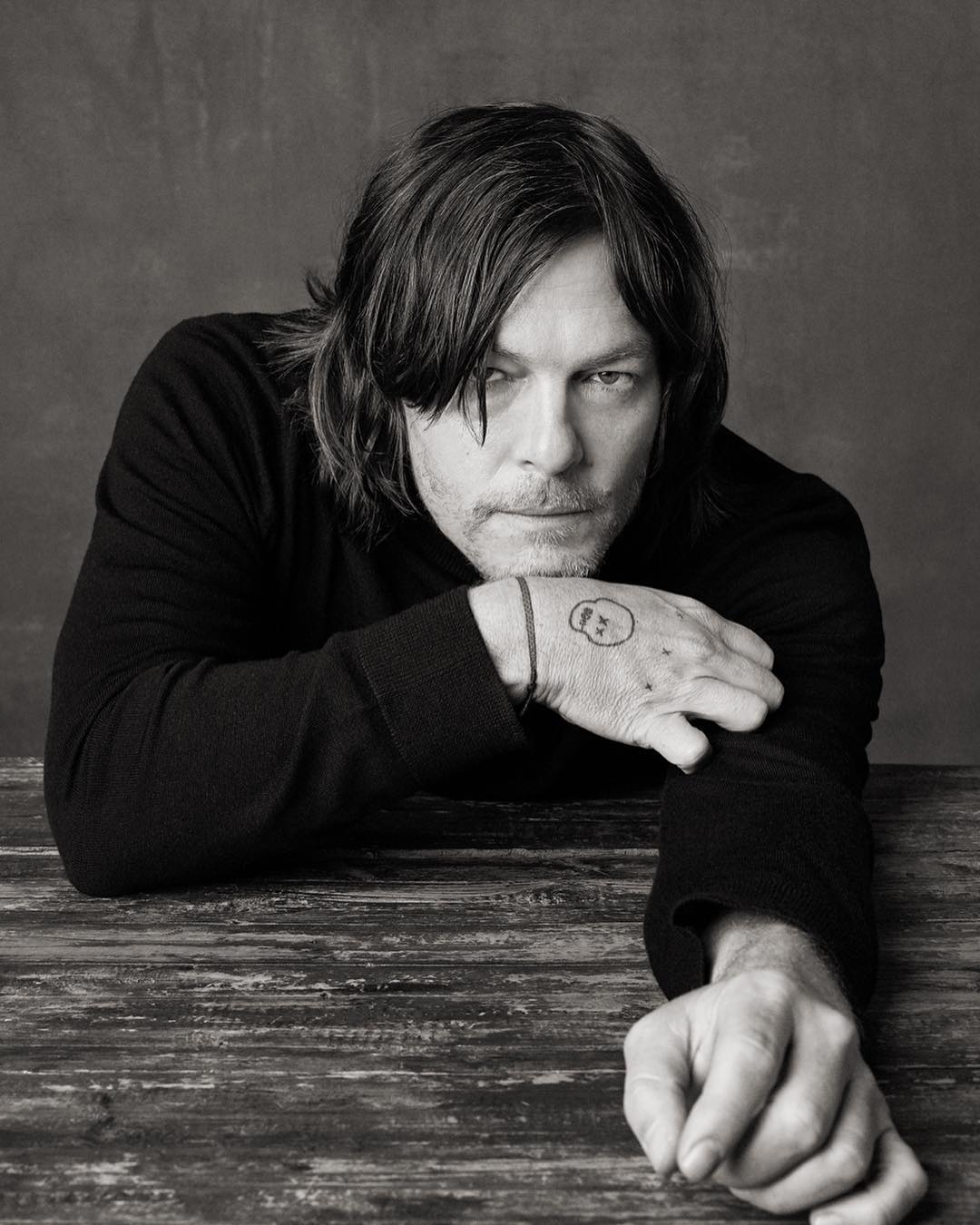 Black and white portrait of Norman Reedus resting his arms on a rustic wooden table, gazing directly at the camera in a photo studio.