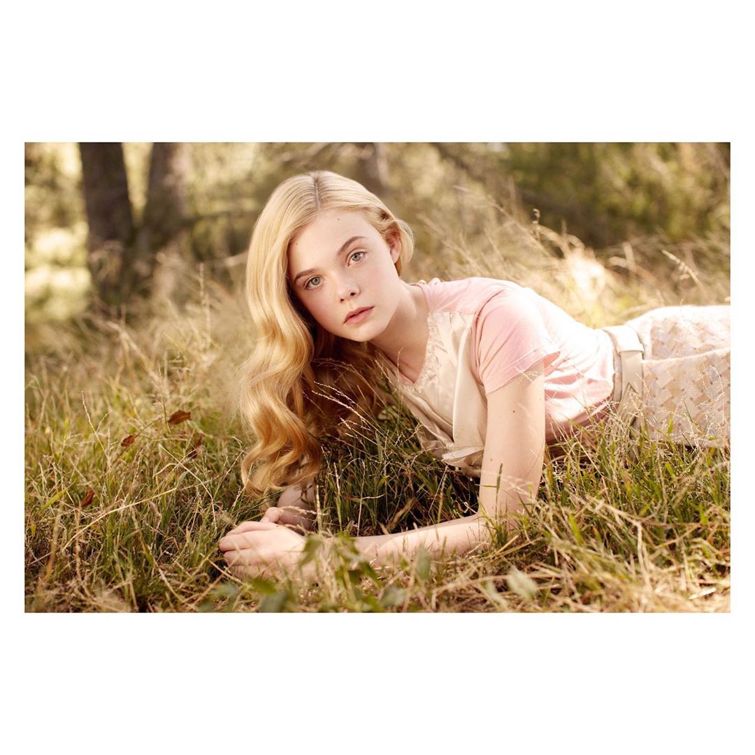 Blonde woman with long hair lying in tall grass wearing a light pink dress in an outdoor, sunlit portrait.