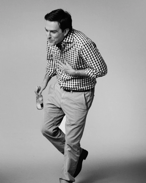 Black and white studio shot of a man in a checkered shirt holding a bottle, with a light gray background and soft lighting.