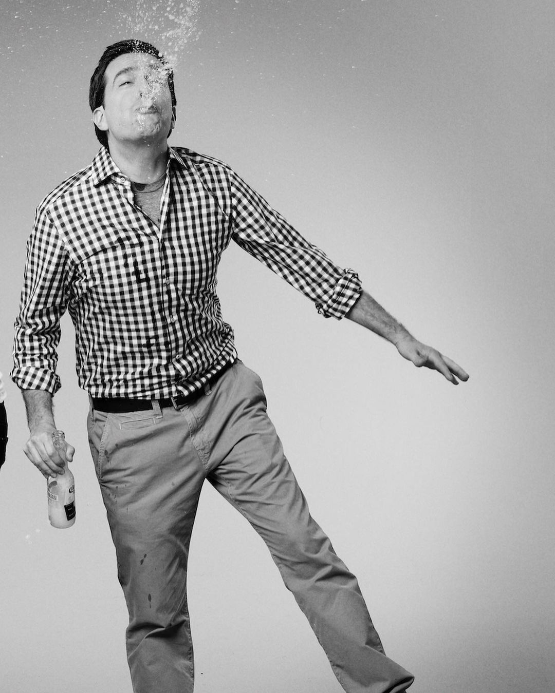 Adult male in casual clothes stands in a photo studio, blowing water from his mouth while holding a bottle, creating a playful, dynamic image.