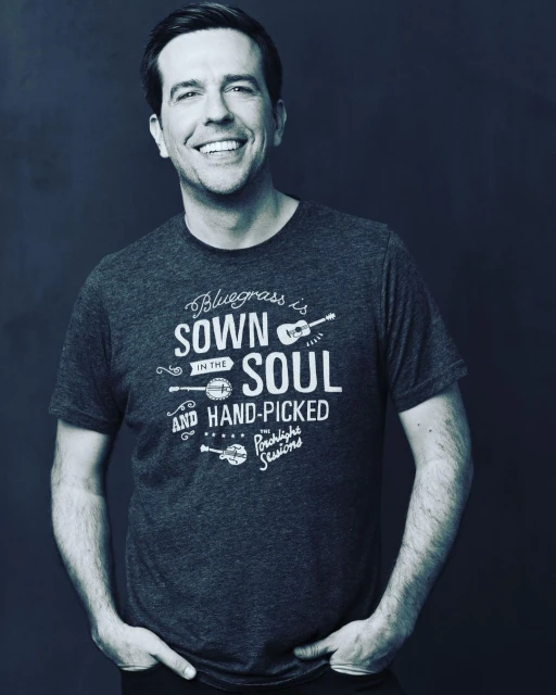 Ed Helms smiles wearing a graphic T-shirt with musical instrument illustrations on a gradient gray background in a medium portrait shot.