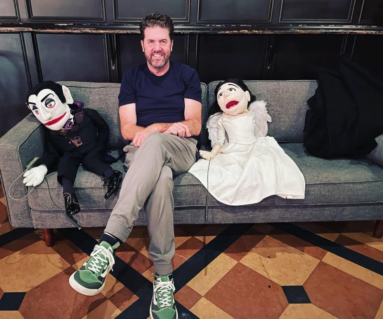 Adult male seated between ventriloquist-style puppets on grey sofa in indoor environment with dark wood-paneled wall behind.