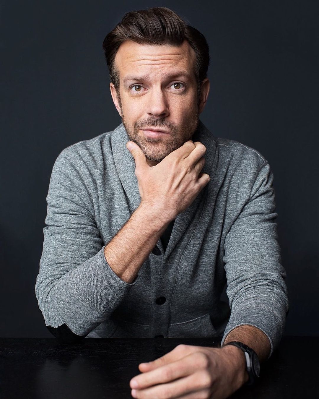 Portrait of a fair-skinned adult male in a grey cardigan, touching his chin, looking at the camera against a dark grey background.