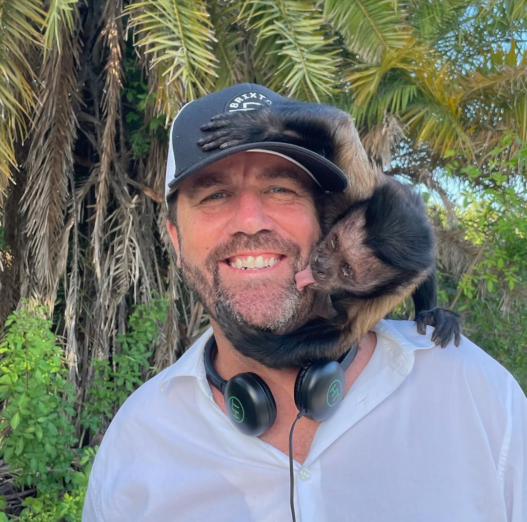 Man in a white shirt and black hat smiles with a brown monkey clinging to his shoulder in an outdoor setting with palm trees.