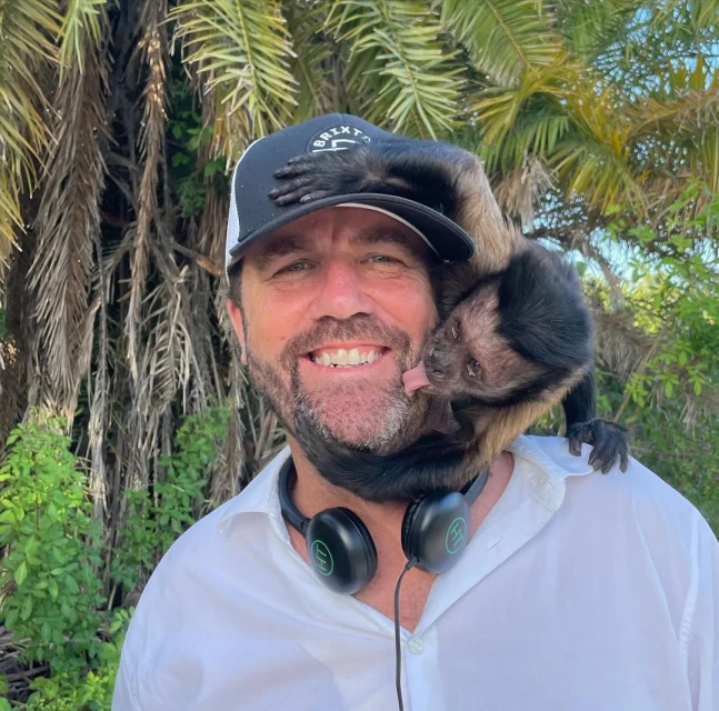 Man in a white shirt and black hat smiles with a brown monkey clinging to his shoulder in an outdoor setting with palm trees.