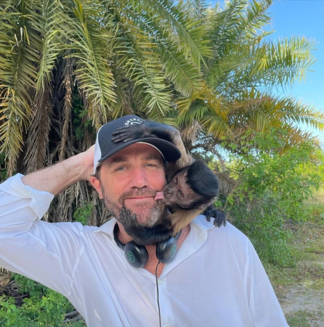 Man poses outdoors with a capuchin monkey on his shoulder; the monkey licks the man's face. The scene includes a palm tree.