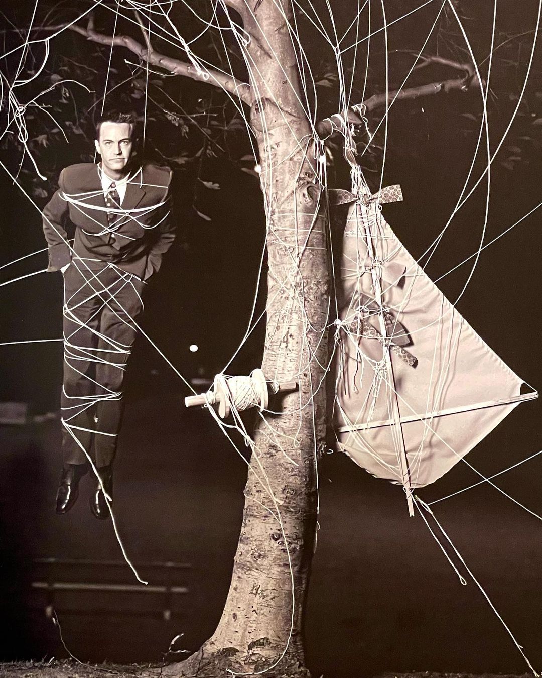 A man in a suit stands tied to a tree with kite string next to a kite in a monochromatic outdoor photograph.