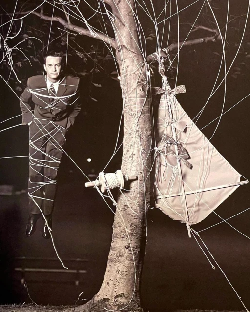 A man in a suit stands tied to a tree with kite string next to a kite in a monochromatic outdoor photograph.