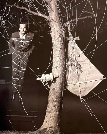 A man in a suit stands tied to a tree with kite string next to a kite in a monochromatic outdoor photograph.
